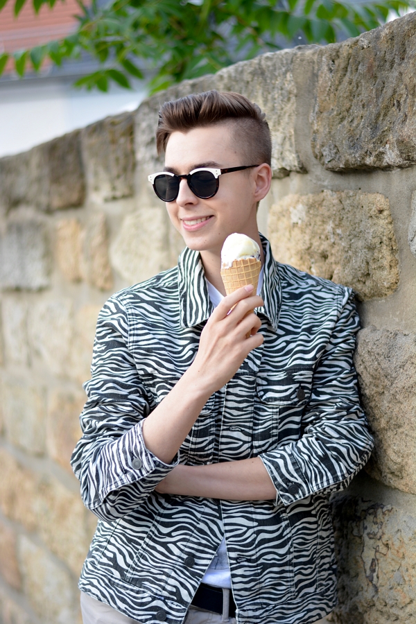 Zebra-print Jeansjacke und schwarz, weiße Sonnenbrille. Modeblogger lacht und genießt Eis. Lehnt an Mauer.